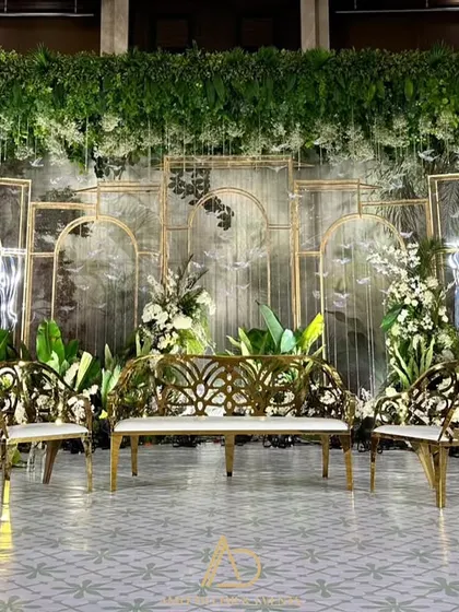 A beautiful stage setup blending modern and natural elements, with a backdrop of artistic panels, lush greenery, and elegant gold and white furniture.