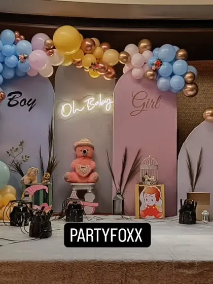 A "Boy or Girl" gender reveal party setup with pink and blue arched backdrops, a central "Oh Baby" neon sign, and a teddy bear prop.