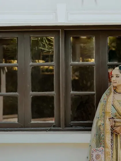 A bride in a unique pastel lehenga stands by a window. This portrait captures her serene beauty and the elegance of her attire in a simple, beautiful composition.