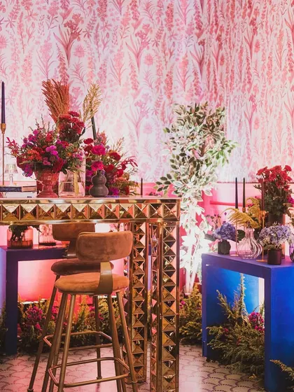 The contrast of the bold blue console tables against the pink patterned wall and red florals creates a vibrant and eclectic look for the sangeet.