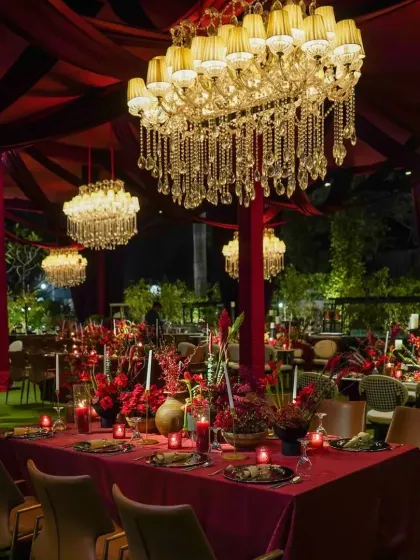 The dining setup combined long tables with rich red linens, deep red floral arrangements, and candlelight to create a dramatic and intimate atmosphere under the hanging chandeliers.