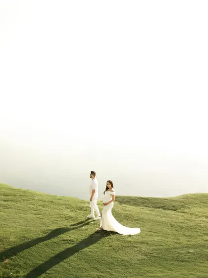A stunning wide-angle shot of the couple on a grassy hill overlooking the water. The vast negative space and golden-hour shadows create a dramatic and minimalist composition.
