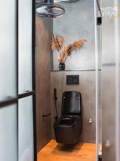 The wet area of the bathroom, featuring a wall-mounted toilet and a rain shower head. The use of dried pampas grass as decor softens the industrial look and adds an organic element.