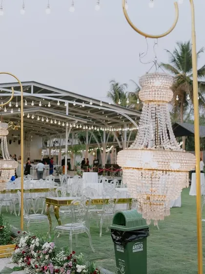 An outdoor lounge area for a reception, featuring modern transparent chairs and tables. The space is defined by stunning, large-scale chandeliers hanging from golden frames, blending luxury with an open-air setting.