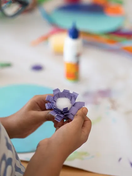 A child's hands carefully assemble a paper flower, showing the delicate process of creating mixed media art. Our workshops encourage kids to explore different materials and textures.