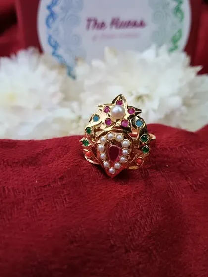 A clear product shot of the "Reva" adjustable ring against a floral background. The design features a teardrop-shaped red stone, pearls, and multi-coloured accents.