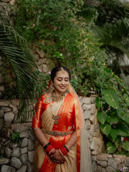 A beautiful outdoor portrait of a bride in a classic cream and red silk saree. The makeup is kept fresh and radiant to complement the natural lighting.