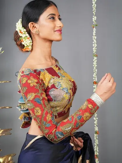 This off-shoulder blouse features long, fitted sleeves with a vibrant red and yellow Kalamkari print. The back has a beautiful tie-up detail with colorful tassels.