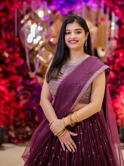 A beautiful portrait of a wedding guest or bridesmaid in a shimmering purple lehenga, smiling against a vibrant red floral wall.