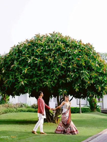 A wide shot of the couple holding hands under a magnificent, sprawling tree, showcasing the natural beauty of their Goan wedding venue.