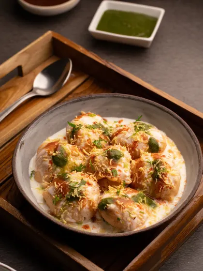 A classic bowl of dahi vada, beautifully garnished and ready to eat. The moody lighting and wooden tray give this traditional snack a sophisticated and inviting feel.