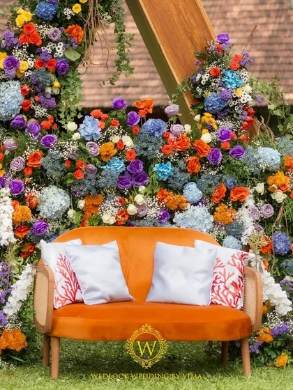 A vibrant floral backdrop and welcome sign for a colorful Haldi and Mehendi ceremony. The mix of orange, blue, purple, and yellow flowers created a stunning photo opportunity for the couple and their guests.