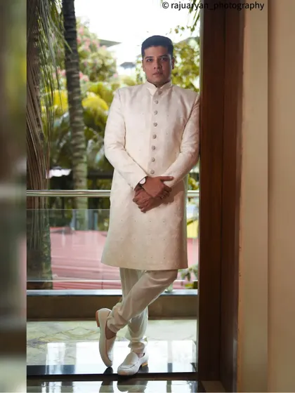 A sharp and stylish full-length portrait of a groom in a classic white sherwani. The natural light from the window and the confident pose create a sophisticated and timeless look for his wedding day.