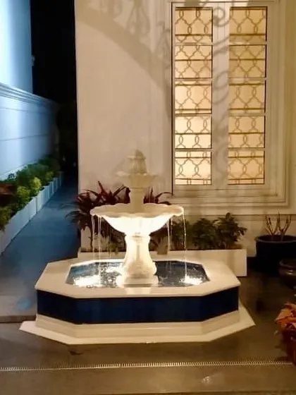 A Moroccan style villa garden featuring a beautiful three tiered white marble fountain as its centerpiece. The fountain is set in an octagonal base with blue tile accents.