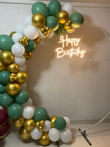 This modern and chic birthday setup features a half-arch of sage green, white, and gold balloons on a simple ring stand. The 'Happy Birthday' neon sign in the center completes the look.