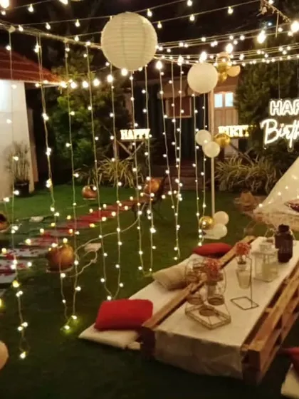 A full view of an elaborate evening garden party. The space is lit by curtains of fairy lights and paper lanterns, with multiple picnic tables and a decorated teepee.