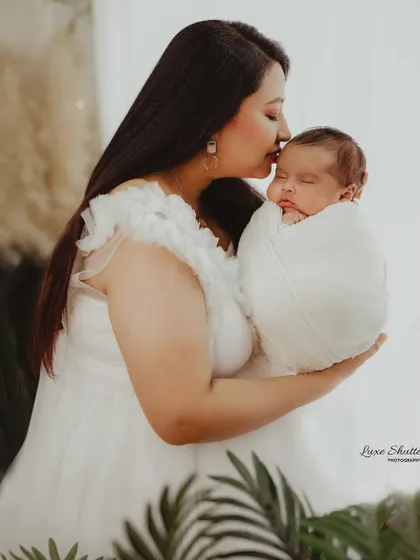 A mother in a white dress gently kissing her swaddled newborn's forehead. The natural elements in the background add a soft, organic feel.