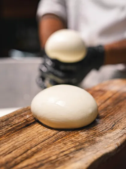 A perfect, glistening ball of fresh mozzarella resting on a wooden board. This is the cheese we use on our pizzas, in our salads, and sell at our deli counters.