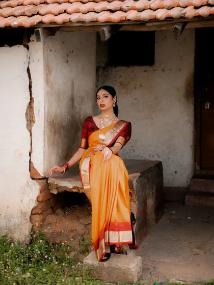 A full-length portrait showing the bride seated on a rustic stone bench. The simplicity of the background makes her vibrant orange saree the hero of the image.