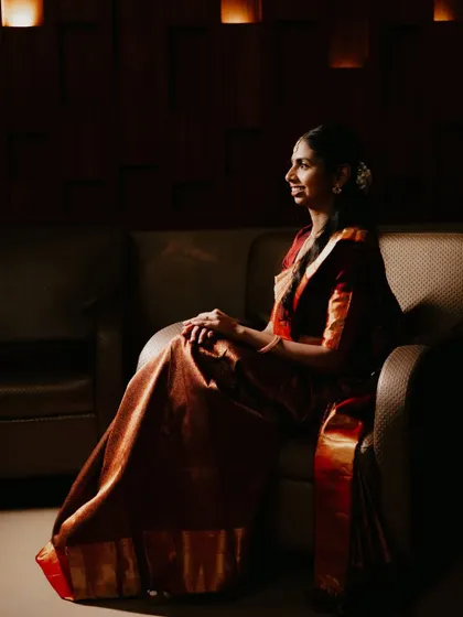 A beautifully lit portrait of the bride seated, her traditional red and gold saree glowing in the warm light.