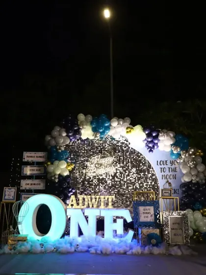 A "Love You to the Moon & Back" themed first birthday setup. The decor features a silver sequin backdrop, a large light-up "ONE" sign, and a blue and silver balloon arch.