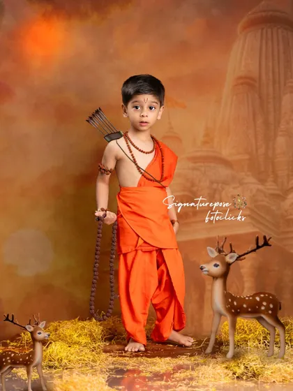 A powerful stance from a little Rama, ready with his bow and arrow. This full-length portrait captures a sense of strength and purpose, with a dramatic, warm-toned backdrop and forest-floor styling.