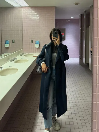 A mirror selfie showing a classic college look with a trench coat and jeans.