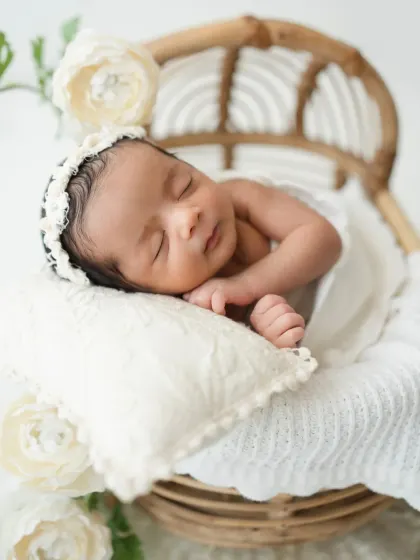 A close-up shot of a newborn girl sleeping peacefully in a cane chair, highlighting her delicate features and the soft, white textures.
