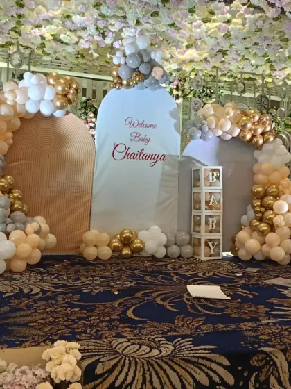 For grand celebrations in large halls, I design elaborate stage setups like this one for baby Chaitanya's welcome party. This features multiple backdrops, large organic balloon arches in gold, white, and beige, and transparent 'PARTY' boxes, all under a stunning floral ceiling.