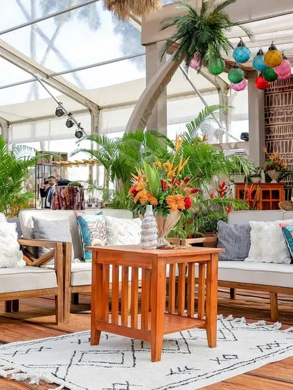 A comfortable lounge area with wooden sofas, Tiki-themed cushions, and a rustic woven rug. The space is surrounded by palm trees and tropical plants, enhancing the island vibe.