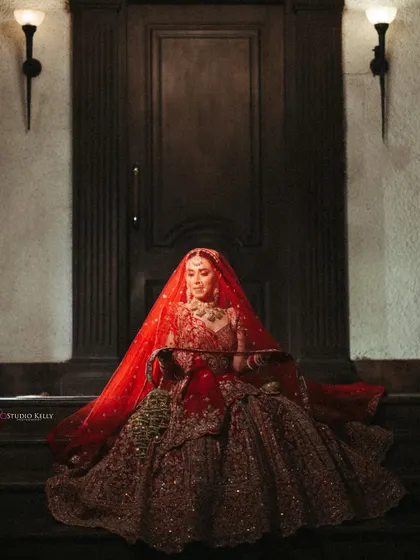 A dramatic, low-light portrait of the bride seated in her full red lehenga, looking regal and timeless.