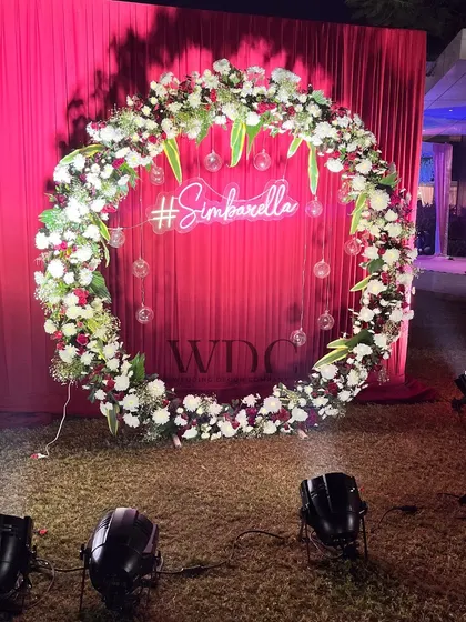 A fun and personal photo-op for the couple. A large floral wreath frames a custom neon sign with their wedding hashtag, set against a backdrop of rich red drapes.