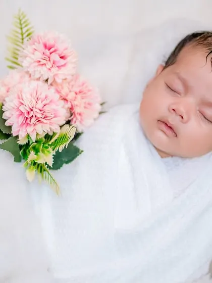 A sweet and simple portrait where the baby appears to be holding a small bouquet of pink carnations. This gentle touch adds a bit of color and charm to a classic white-on-white newborn photo.