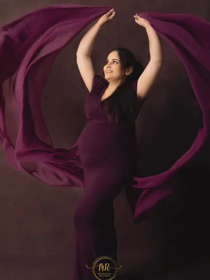 A joyful and dynamic solo portrait. The mother-to-be is wearing a deep purple gown, tossing the fabric in the air to create a beautiful, flowing shape around her.