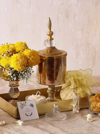 The Sweet Magnolia hamper is a complete festive set. It includes a candle, a potli with trail mix, a flower vase with marigolds, and a glass jar of mini cookies on a golden metal tray.