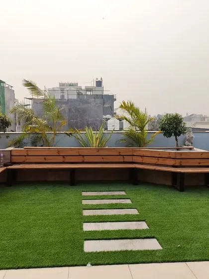 A custom-built L-shaped bench made from thermopine wood, set on an artificial lawn with a stone pathway. The bench is integrated with large planters.