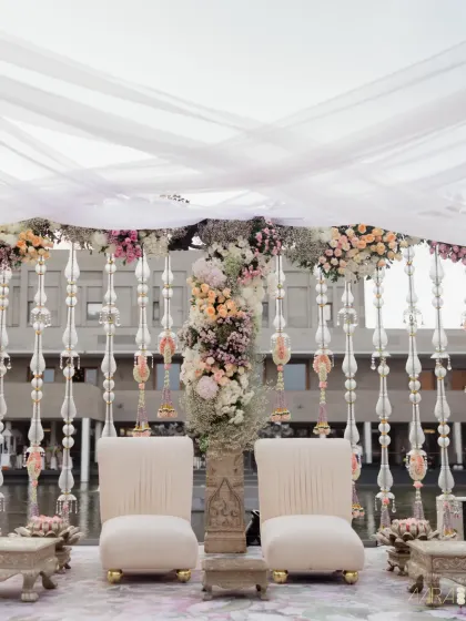 The wedding couple's seating at a breezy, waterside engagement. The elegant white chairs are framed by hanging crystal tassels and lush pastel floral arrangements, creating a picture-perfect focal point.