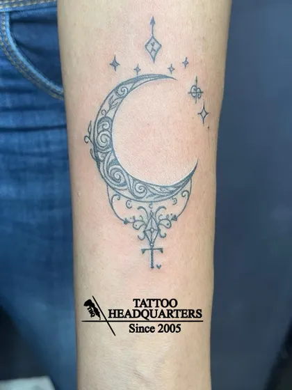 An ornamental crescent moon tattoo on the forearm, featuring intricate swirls and sparkling star accents for a magical look.