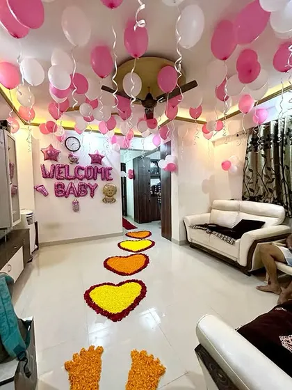 A heartwarming welcome home decoration for a newborn baby. The setup shows a path of marigold flower hearts leading into the living room, with pink and white balloons on the ceiling and "WELCOME BABY" foil balloons on the wall.