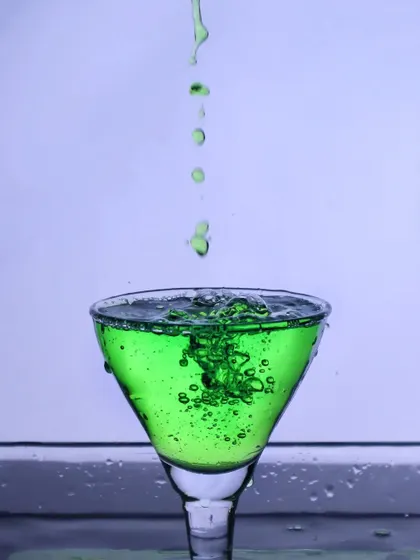 A vibrant green drink being poured into a glass, creating bubbles and motion. The clean, simple background makes the color pop, resulting in a refreshing and eye-catching shot.