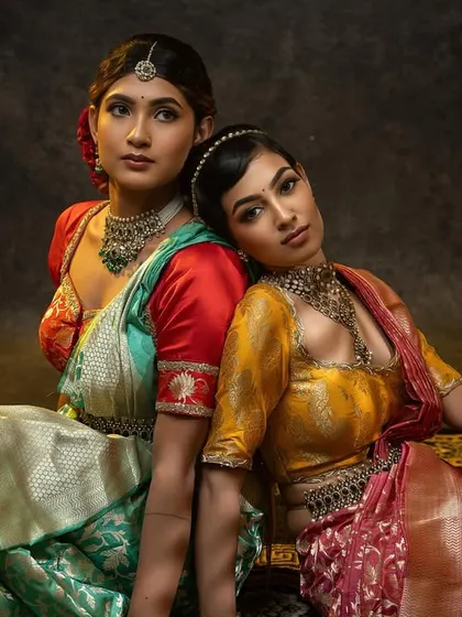 Two models showcasing our custom blouses. One wears a red blouse with a green saree, the other a yellow blouse with a pink saree, both featuring our signature embroidery.