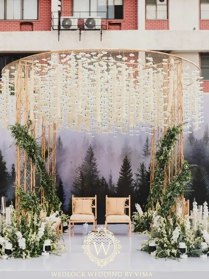 A modern minimalist mandap that perfectly blends nature and contemporary design. The structure features a backdrop of a misty forest, a canopy of hanging white florals, and lush green arrangements.