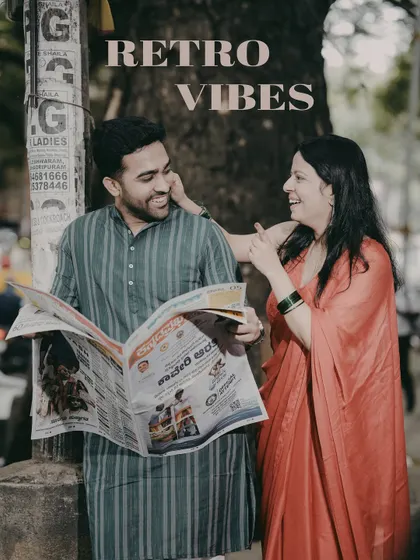 A couple shares a laugh while reading a newspaper, creating a candid and charming retro-themed pre-wedding photo on a city street.