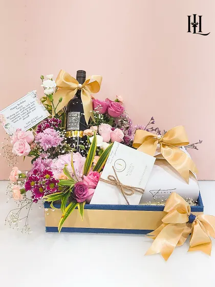 This birthday hamper combines elegance and indulgence. A bottle of fine wine is nestled amongst a lush arrangement of fresh flowers, accompanied by a spa voucher and other curated gifts, all presented in a stylish blue and gold tray.