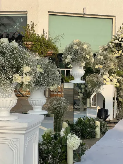 The pathway to the wedding is adorned with arrangements of baby's breath and white roses in classic urns, set on mirrored pedestals that reflect the beauty.