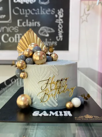 A sophisticated grey birthday cake for Samir, featuring a delicate white leaf stencil pattern. The cake is accented with gold and grey spheres, a dried palm leaf, and a gold "Happy Birthday" topper.