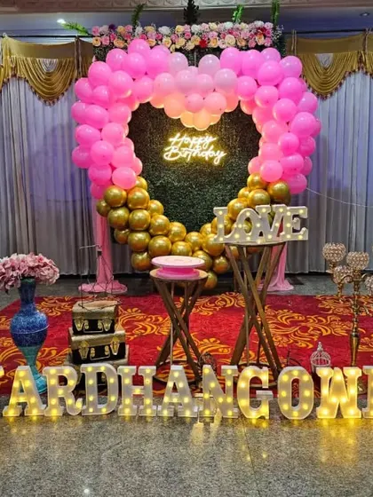 A heart-shaped balloon backdrop in pink and gold for Vardhan's birthday. The setup includes a 'LOVE' sign, marquee letters for the name, and a small picket fence for a charming touch.