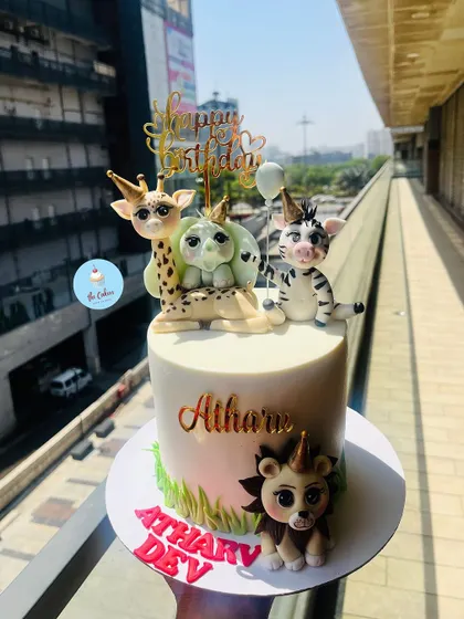 A charming animal party cake with fondant figures of a giraffe, cheetah, and zebra all wearing party hats. A small lion at the base completes this fun celebration scene.