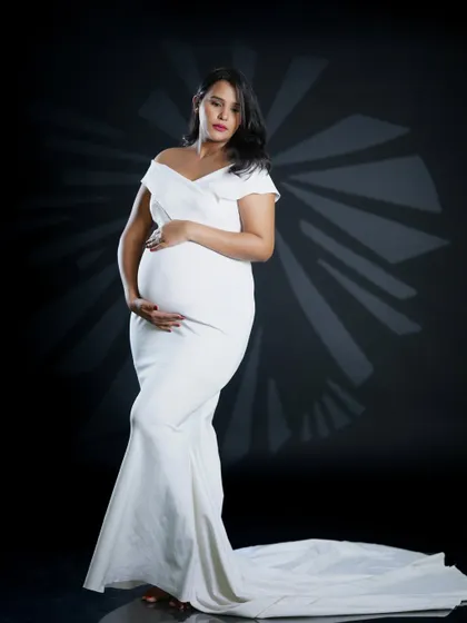 This portrait showcases an elegant, off-the-shoulder white gown that beautifully drapes over the baby bump. The dramatic studio lighting creates a sophisticated and timeless look, focusing entirely on the mother-to-be.