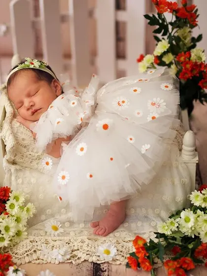 A dreamy garden-themed newborn shoot. The baby is dressed in a daisy-print outfit, sleeping on a miniature bed surrounded by vibrant flowers and a white picket fence.
